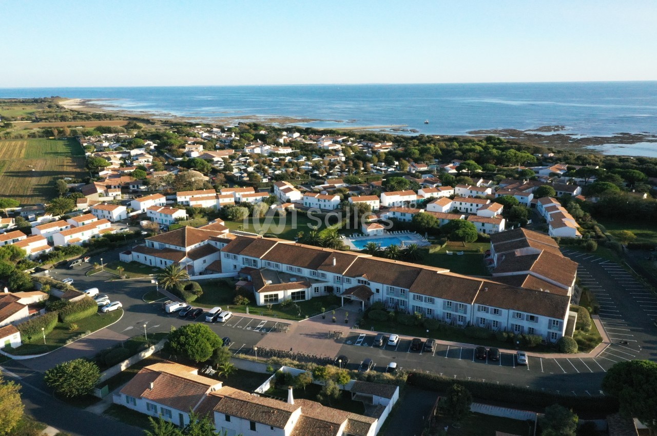 Vue aérienne d'un village côtier avec maisons blanches et toits rouges, entouré de verdure et bordé par la mer.