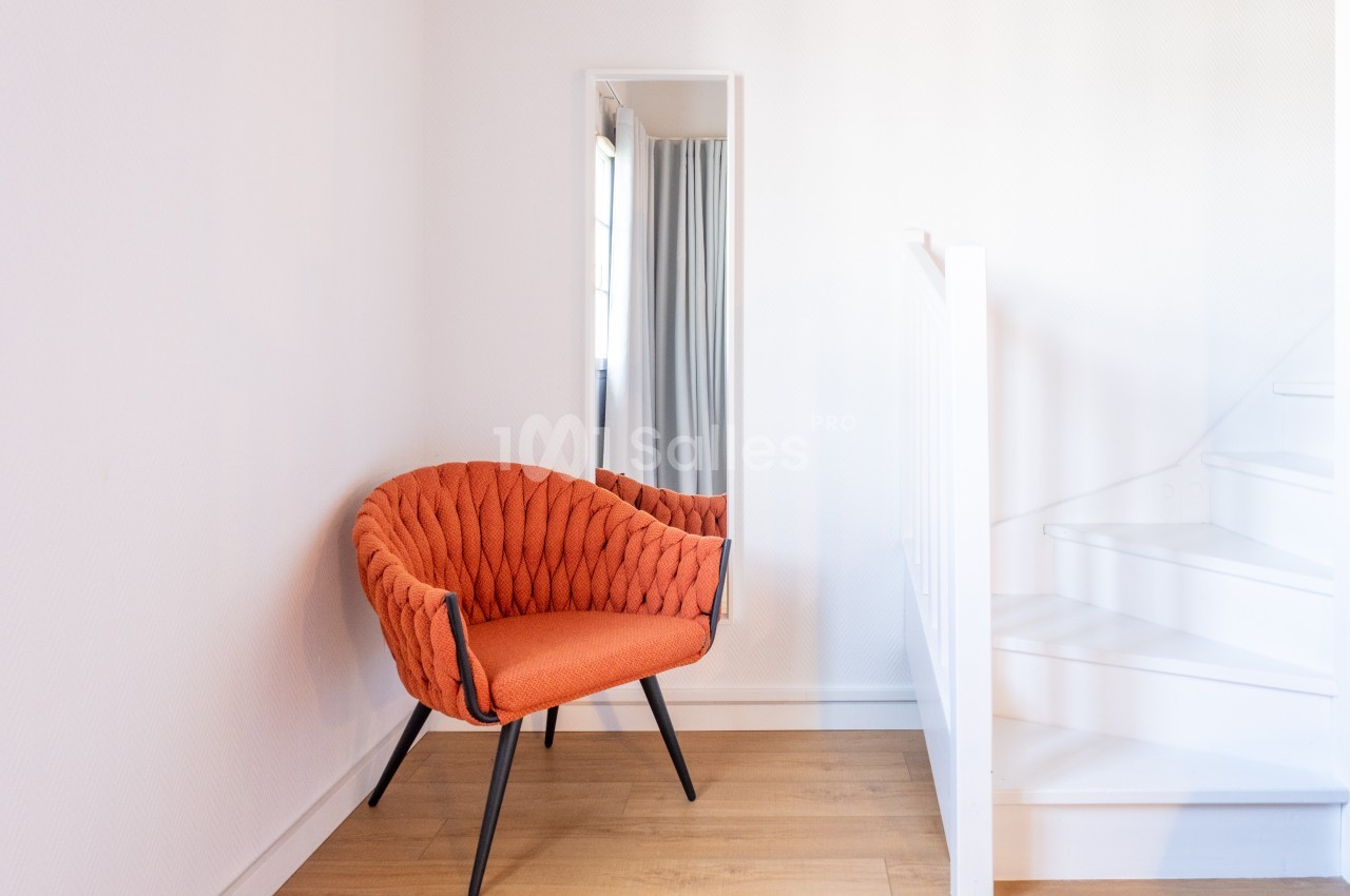 Fauteuil orange au design moderne placé près d'un miroir et d'un escalier blanc dans une pièce lumineuse.