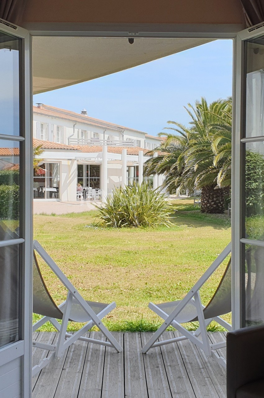 Vue depuis une porte-fenêtre ouverte sur une terrasse en bois, deux chaises et un jardin avec palmiers et bâtiment blanc.