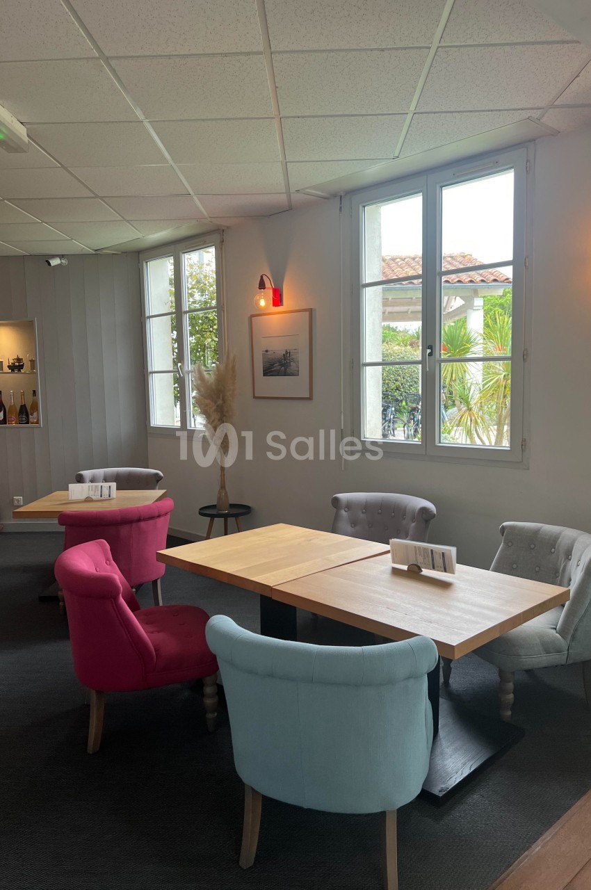 Salle lumineuse avec tables en bois, chaises colorées et décoration sobre, vue sur l'extérieur par de grandes fenêtres.