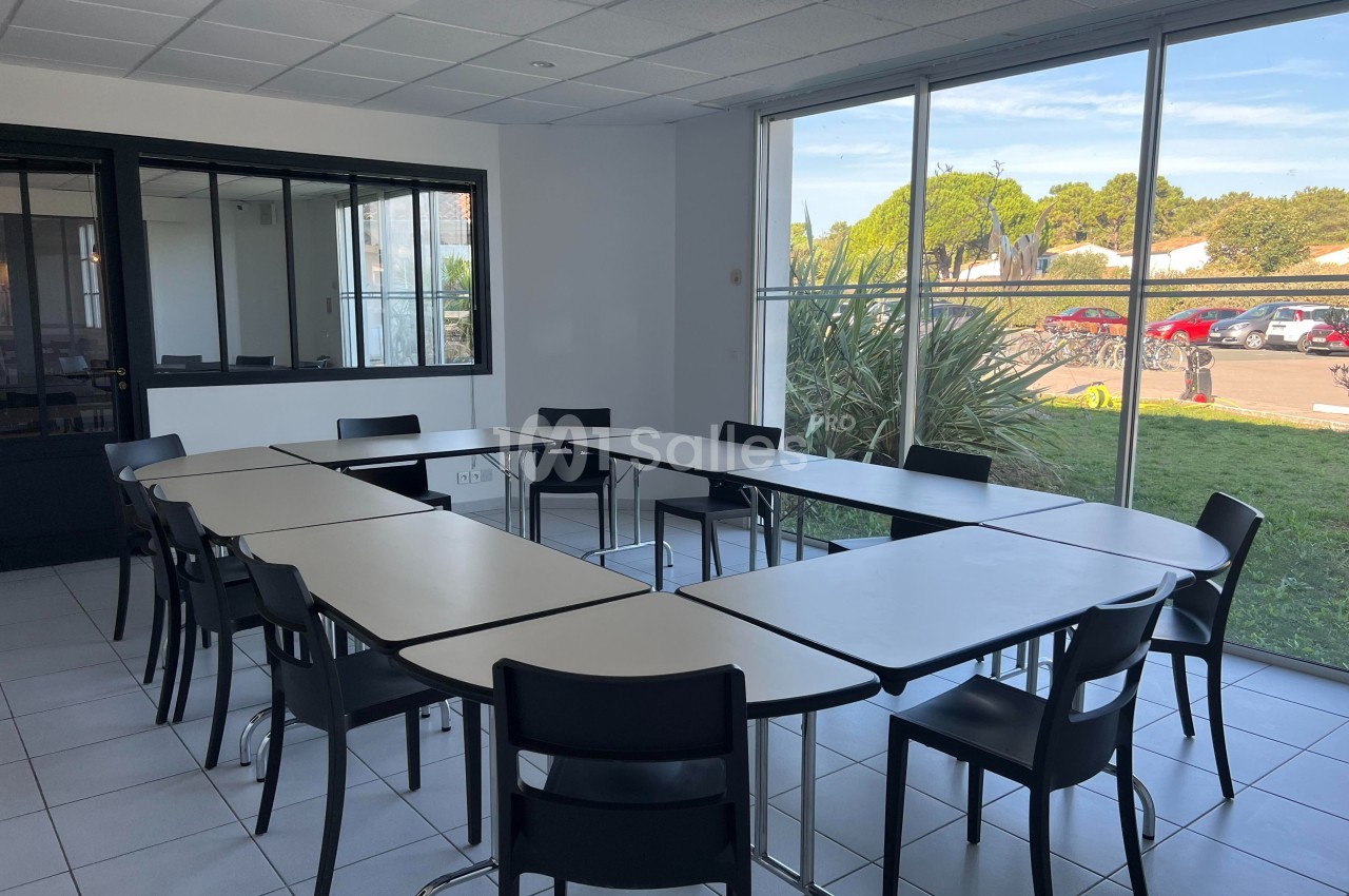 Salle de réunion lumineuse avec tables disposées en U, chaises noires et grande baie vitrée donnant sur un espace extérieur.