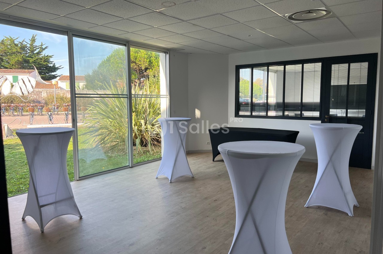 Salle lumineuse avec grandes baies vitrées, tables hautes drapées de blanc et vue sur un espace extérieur verdoyant.