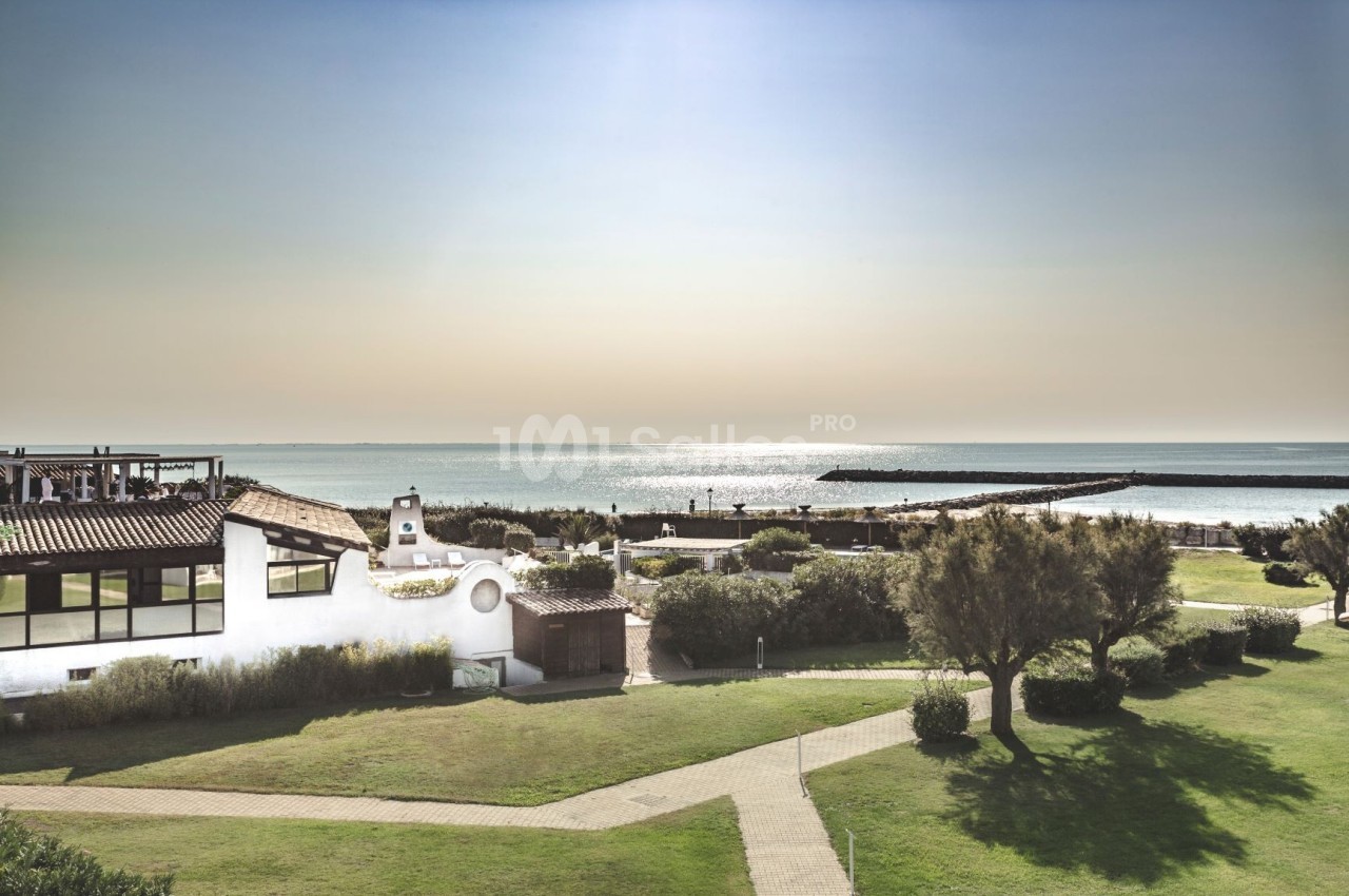 Vue d'un bâtiment blanc entouré de pelouses avec un chemin menant à la mer et une jetée à l'horizon.