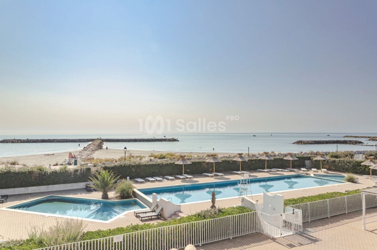 Vue sur une piscine extérieure avec transats, entourée de végétation, donnant sur une plage et la mer calme.
