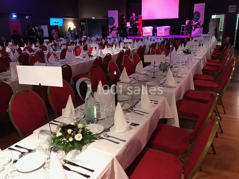 Salle de réception avec tables dressées, nappes blanches, chaises rouges et décorations florales.