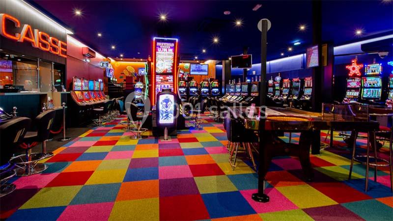 Salle de jeux avec machines à sous, sol coloré en damier, éclairage néon et espace caisse visible à gauche.