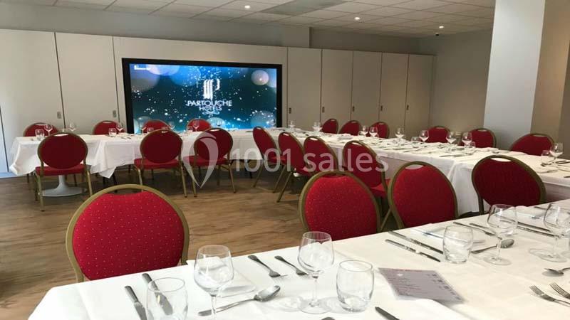 Salle de réception avec tables dressées, nappes blanches, chaises rouges et écran affichant un visuel lumineux.
