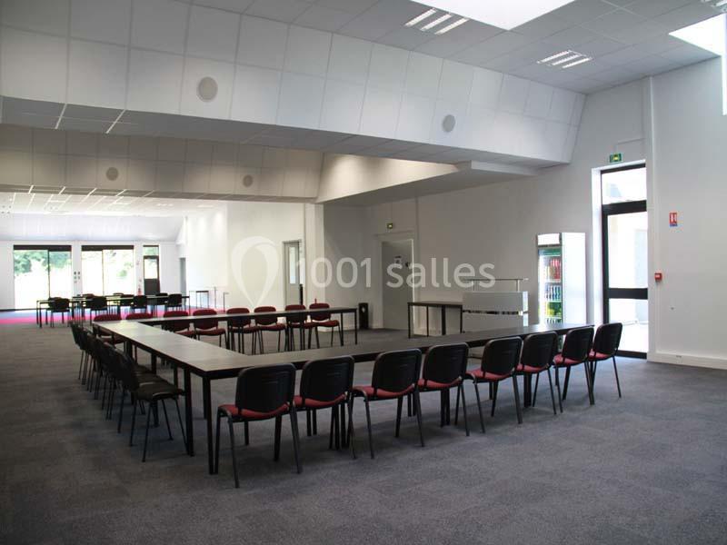 Salle de réunion spacieuse avec des tables disposées en U, des chaises rouges et un éclairage naturel.