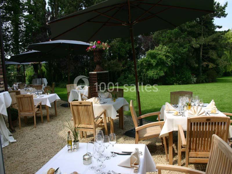 Terrasse d'un restaurant avec tables dressées, chaises en bois, parasols noirs et vue sur un jardin verdoyant.