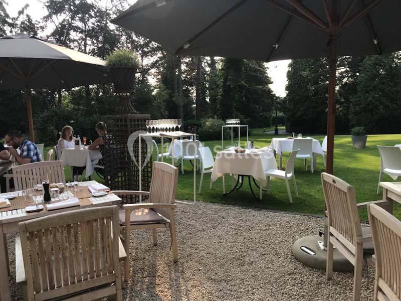 Terrasse extérieure avec tables dressées, chaises blanches et parasols, entourée de pelouse et d'arbres.