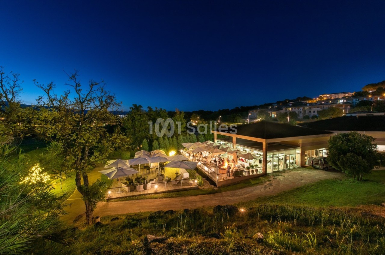 Terrasse éclairée d'un restaurant entourée de végétation, vue de nuit avec un ciel bleu profond.