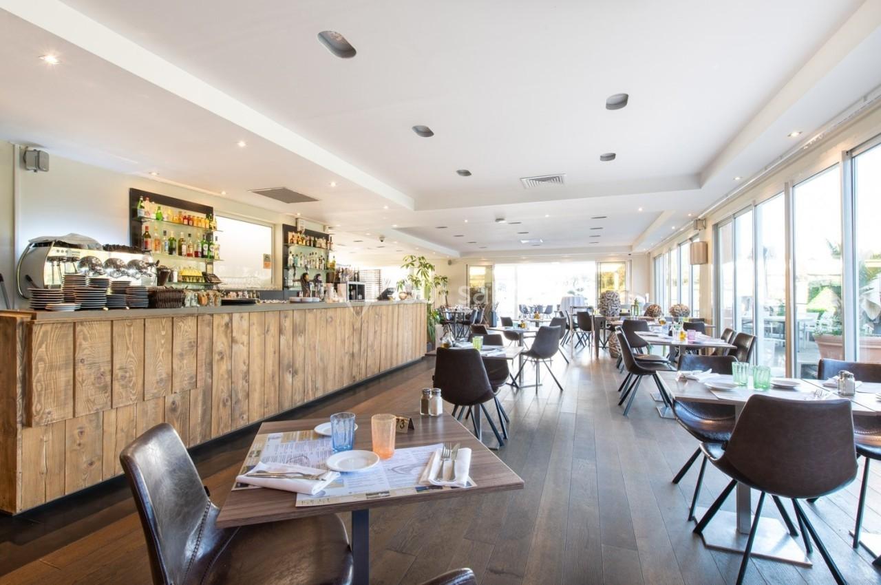 Salle de restaurant lumineuse avec un bar en bois, des tables dressées et de grandes baies vitrées donnant sur l'extérieur.
