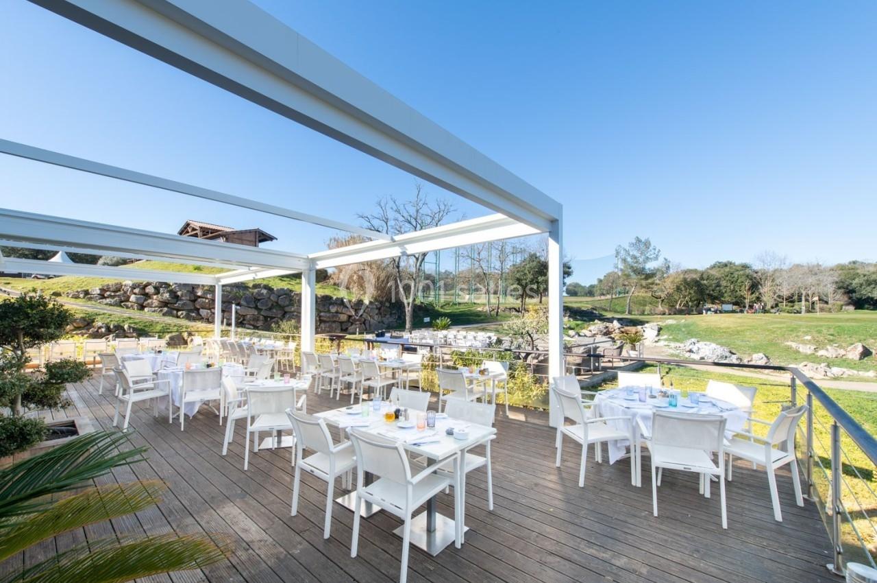 Terrasse en bois aménagée avec des tables et chaises blanches, entourée de verdure et baignée de lumière naturelle.