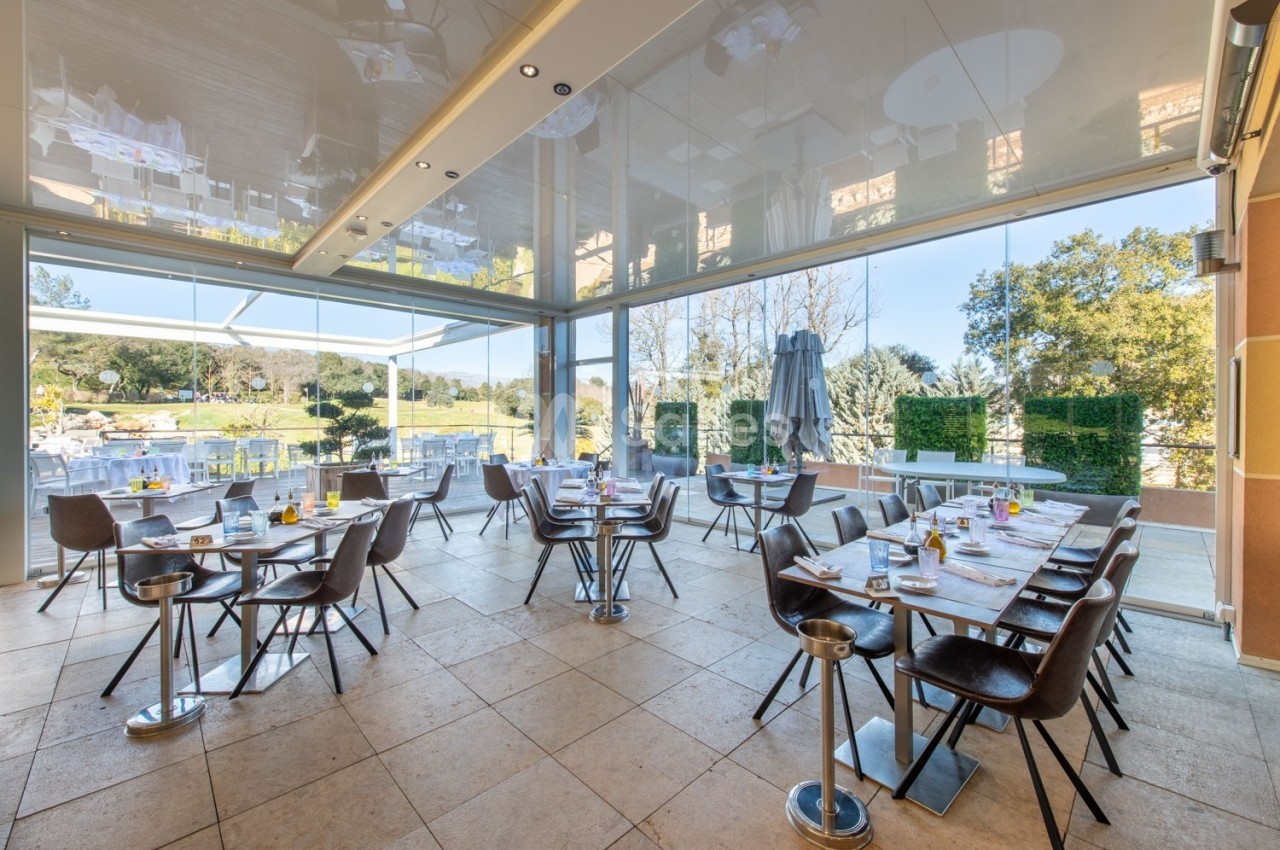 Salle de restaurant lumineuse avec tables dressées, grandes baies vitrées donnant sur un espace extérieur verdoyant.