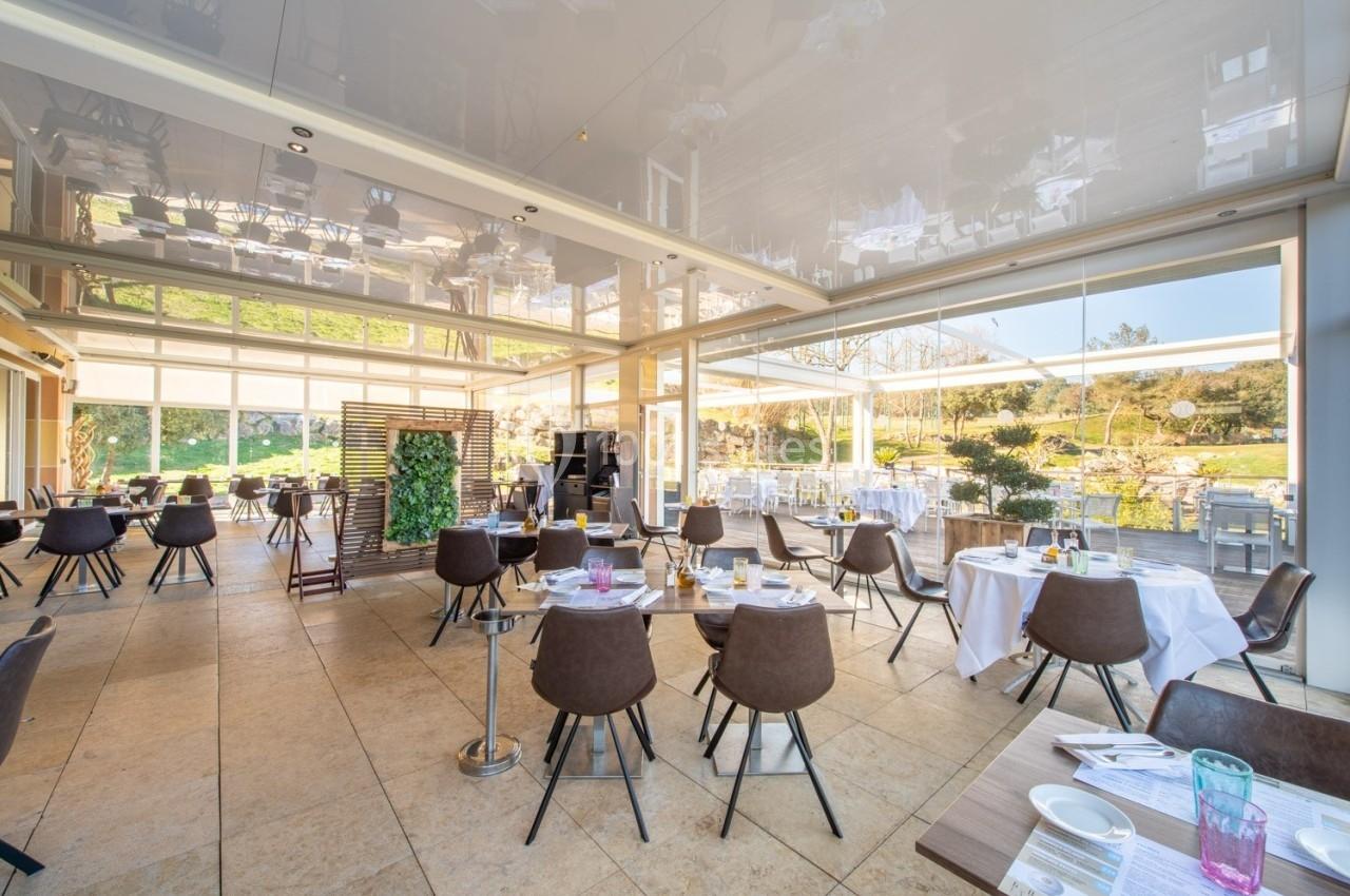 Salle de restaurant lumineuse avec grandes baies vitrées, tables dressées et vue sur un jardin extérieur.