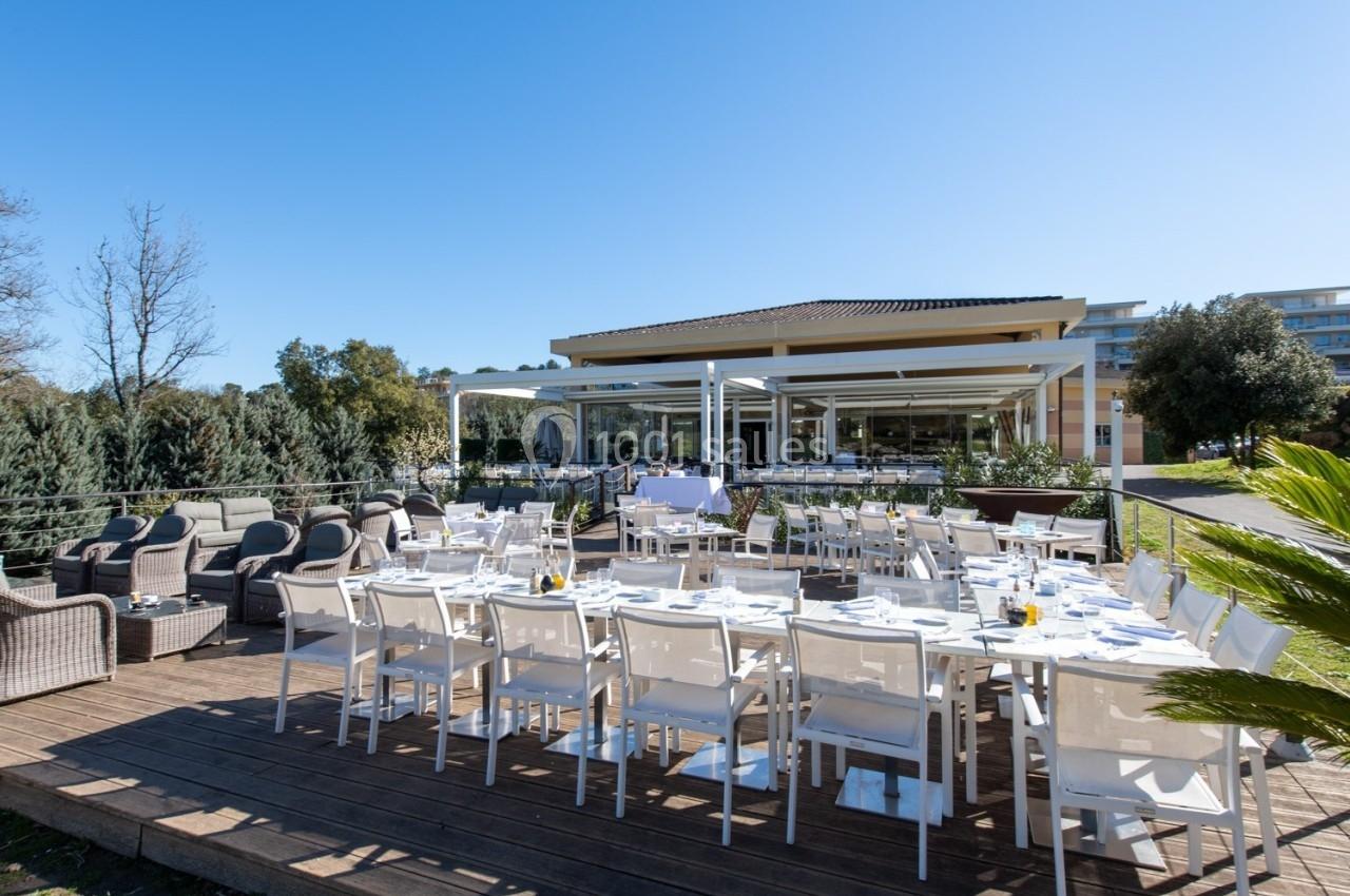 Terrasse aménagée avec tables et chaises blanches, entourée de verdure, devant un bâtiment moderne avec baies vitrées.