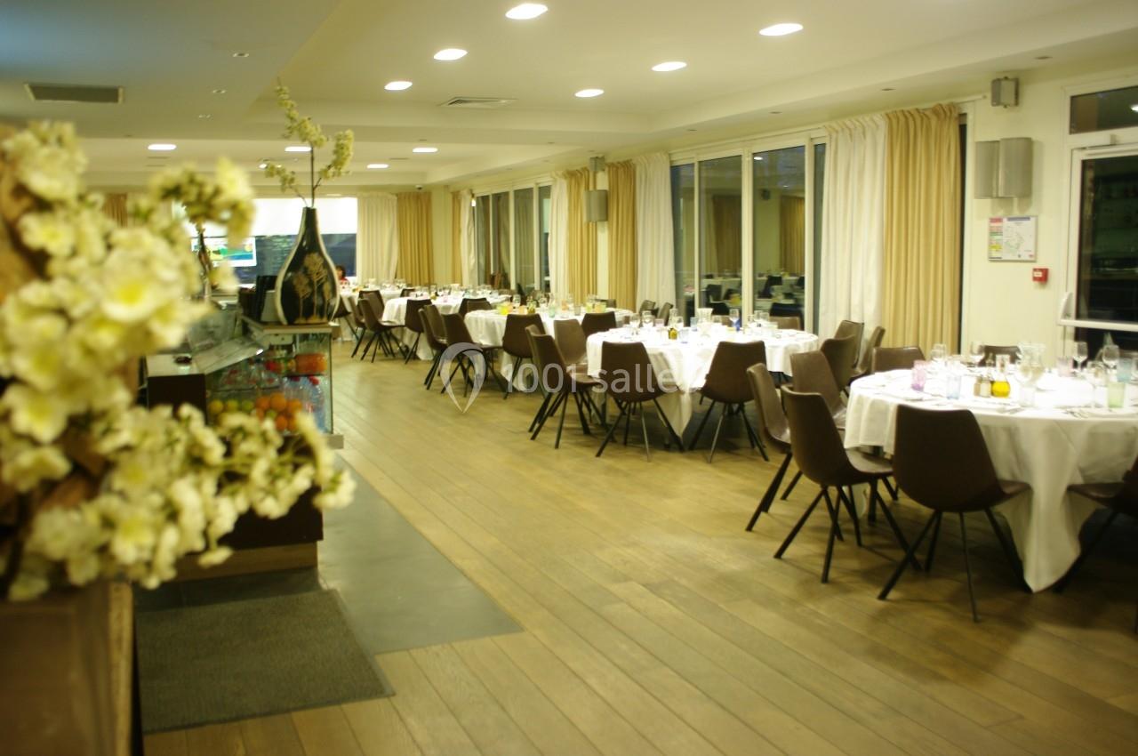 Salle de restaurant lumineuse avec tables dressées, chaises marron et grandes baies vitrées donnant sur l'extérieur.