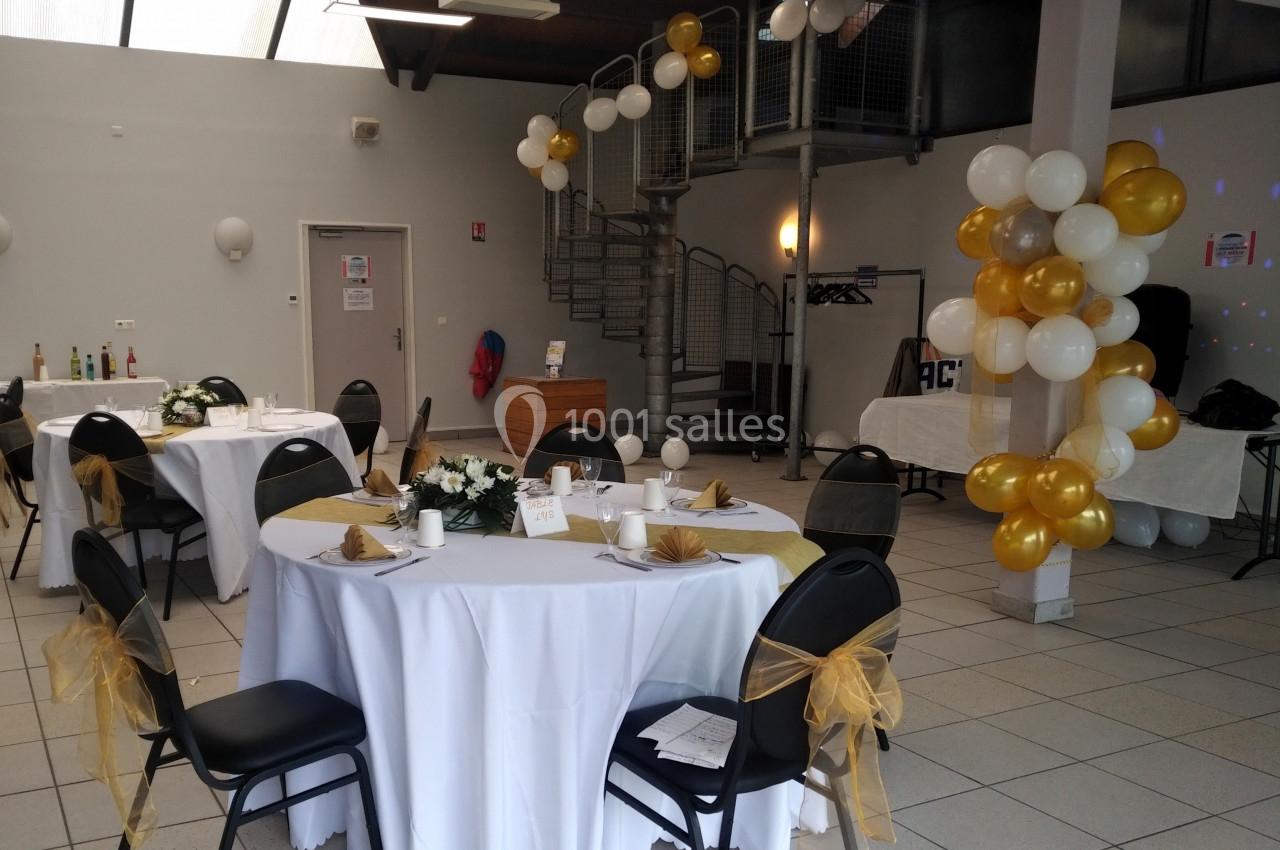 Salle décorée pour un événement avec tables rondes dressées, nappes blanches, chaises ornées et ballons dorés et blancs.