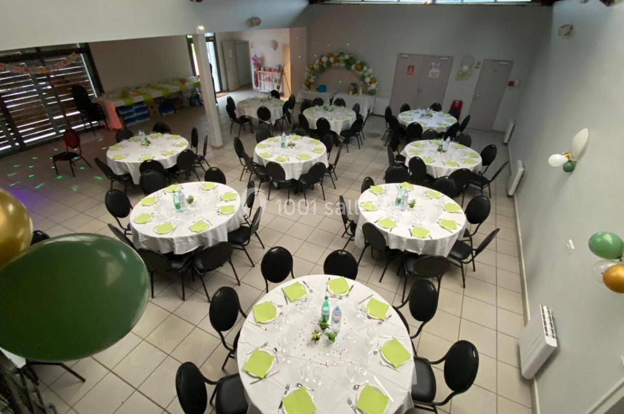 Salle décorée avec des tables rondes dressées, nappes blanches, serviettes vertes et ballons pour un événement festif.
