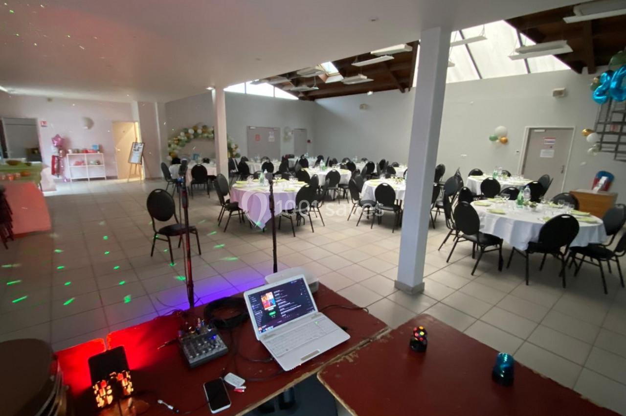 Salle de réception lumineuse avec tables rondes dressées, chaises noires et équipement audio en premier plan.