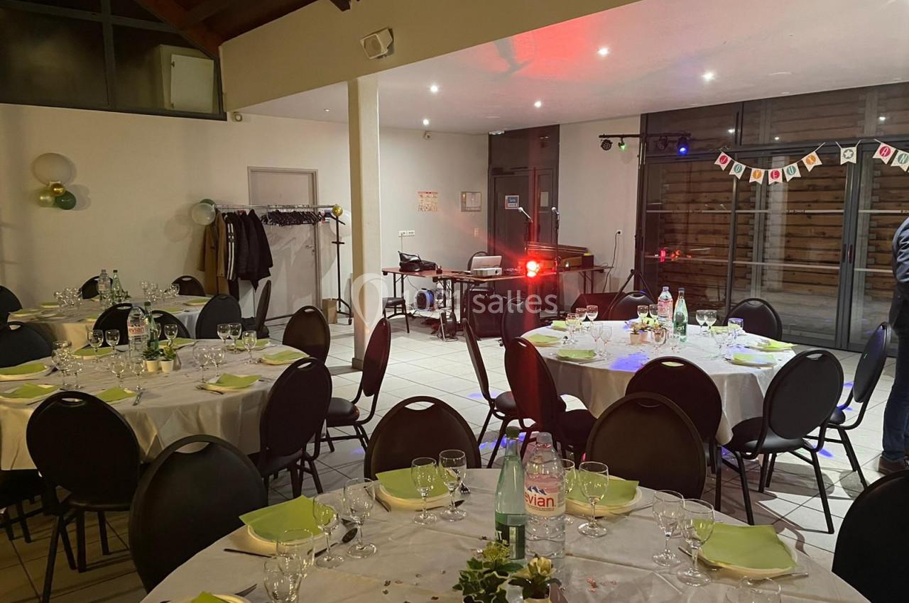 Salle décorée pour un événement, avec tables rondes dressées, chaises noires et éclairage tamisé.