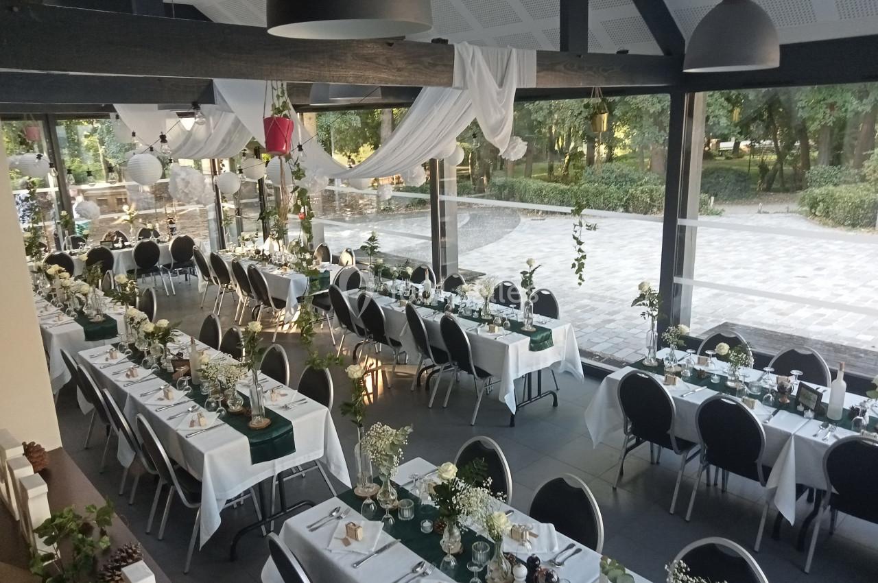 Salle de réception décorée avec des tables alignées, nappes blanches, fleurs et vue sur un jardin extérieur.