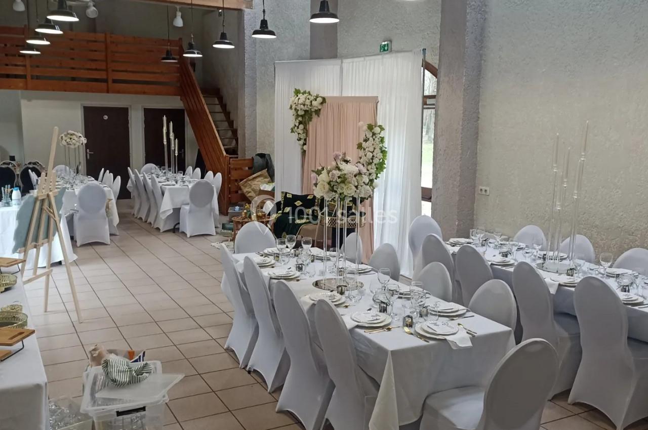 Salle de réception décorée pour un événement, avec des tables dressées, chaises blanches et un espace fleuri.