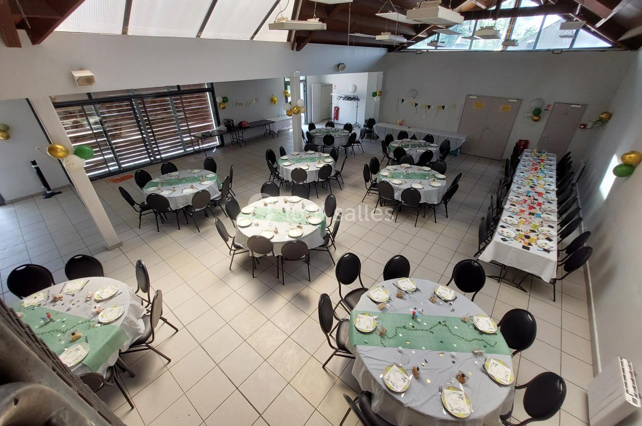 Salle décorée avec des tables rondes et une longue table, préparées pour un événement avec nappes vertes et vaisselle.