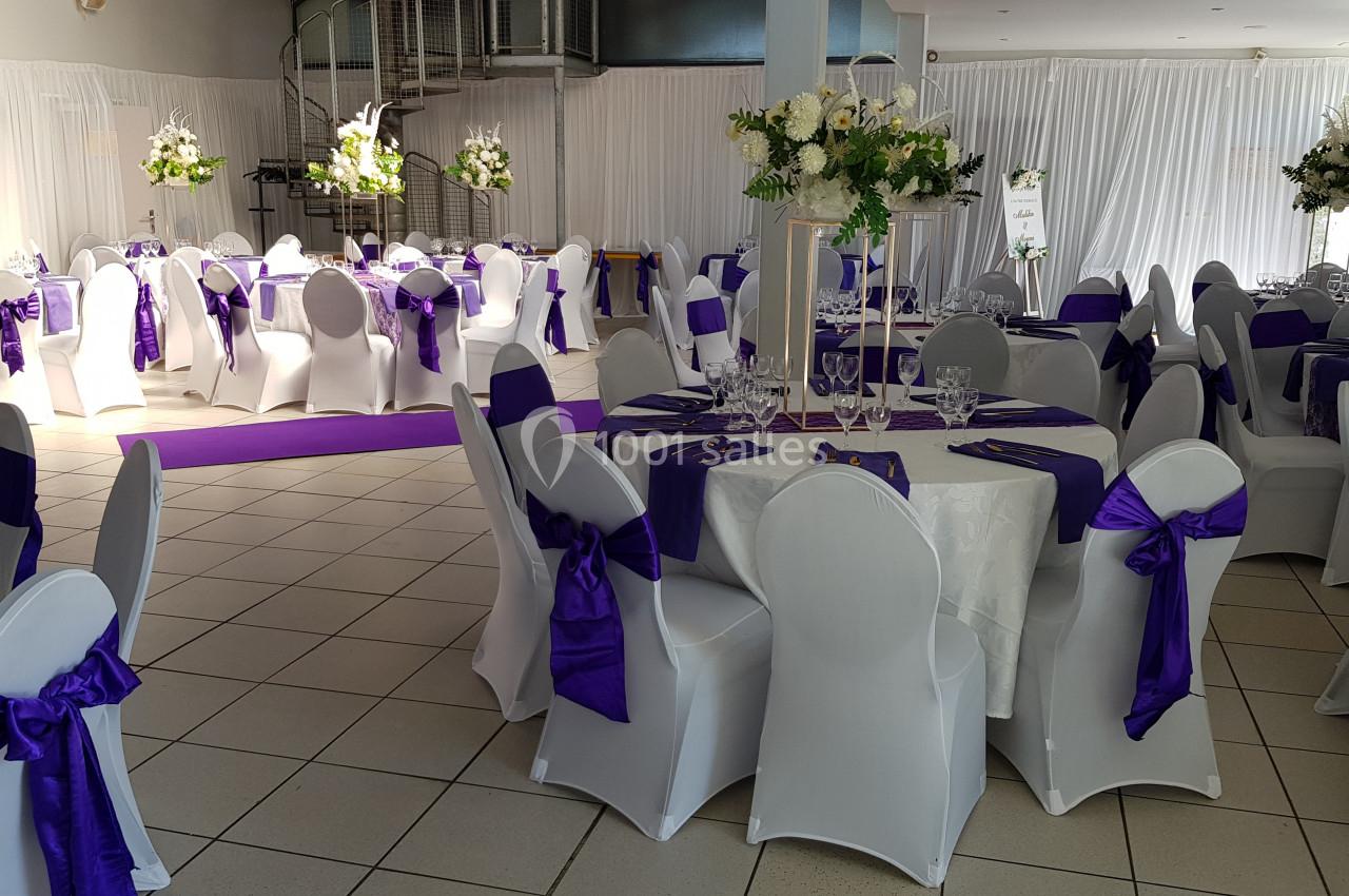 Salle de réception décorée avec des nappes blanches, des chaises ornées de nœuds violets et des arrangements floraux.