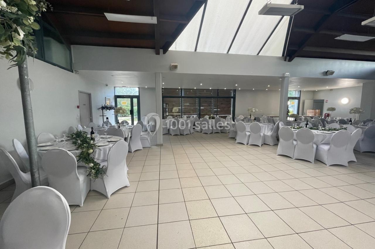 Salle de réception lumineuse avec tables rondes dressées, chaises blanches et décoration florale.