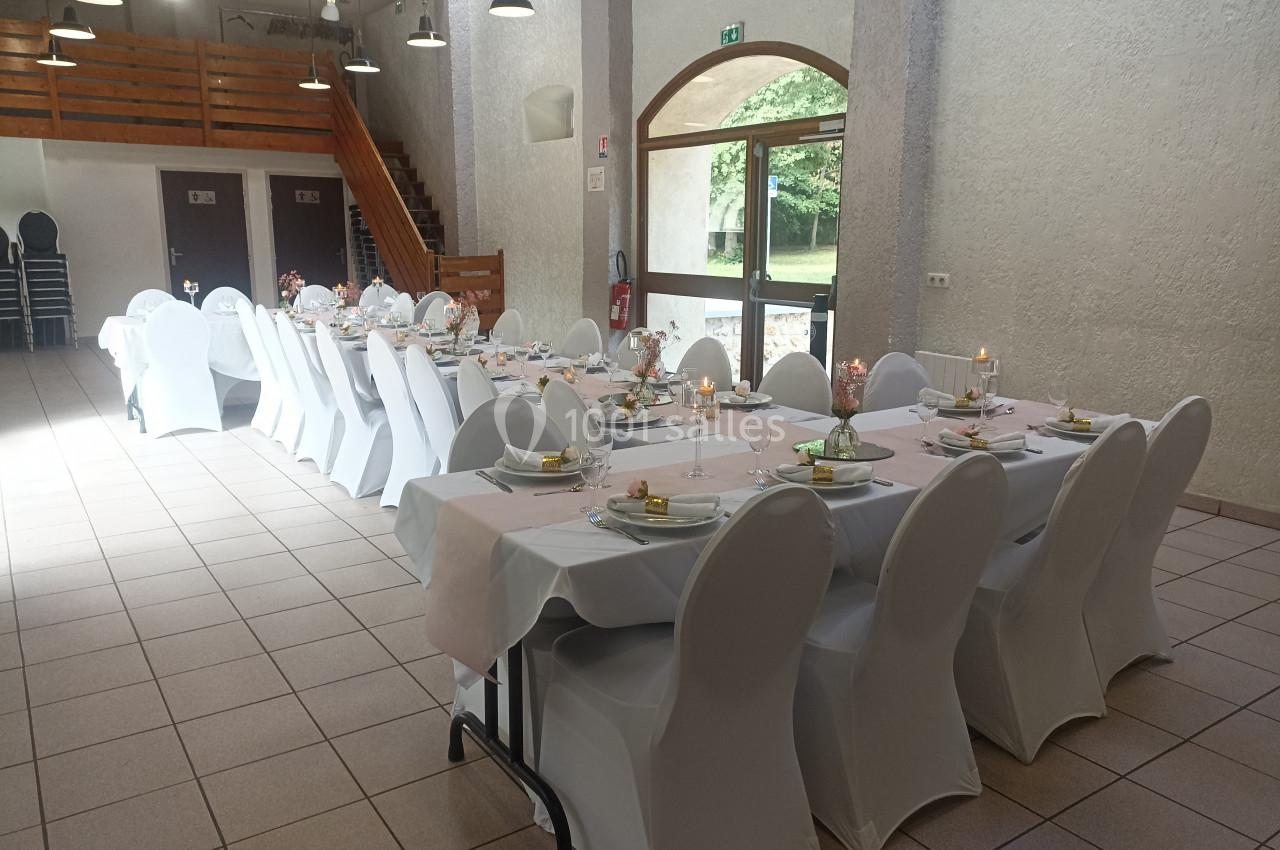 Salle décorée pour un repas, avec tables alignées, nappes blanches, chaises houssées et lumière naturelle entrant par une…