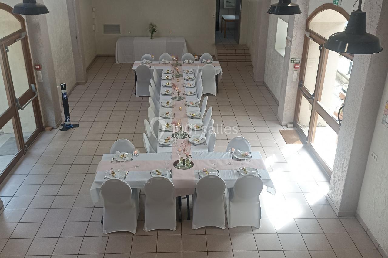 Salle lumineuse avec une grande table dressée pour un repas, entourée de chaises blanches, sous des lampes suspendues.