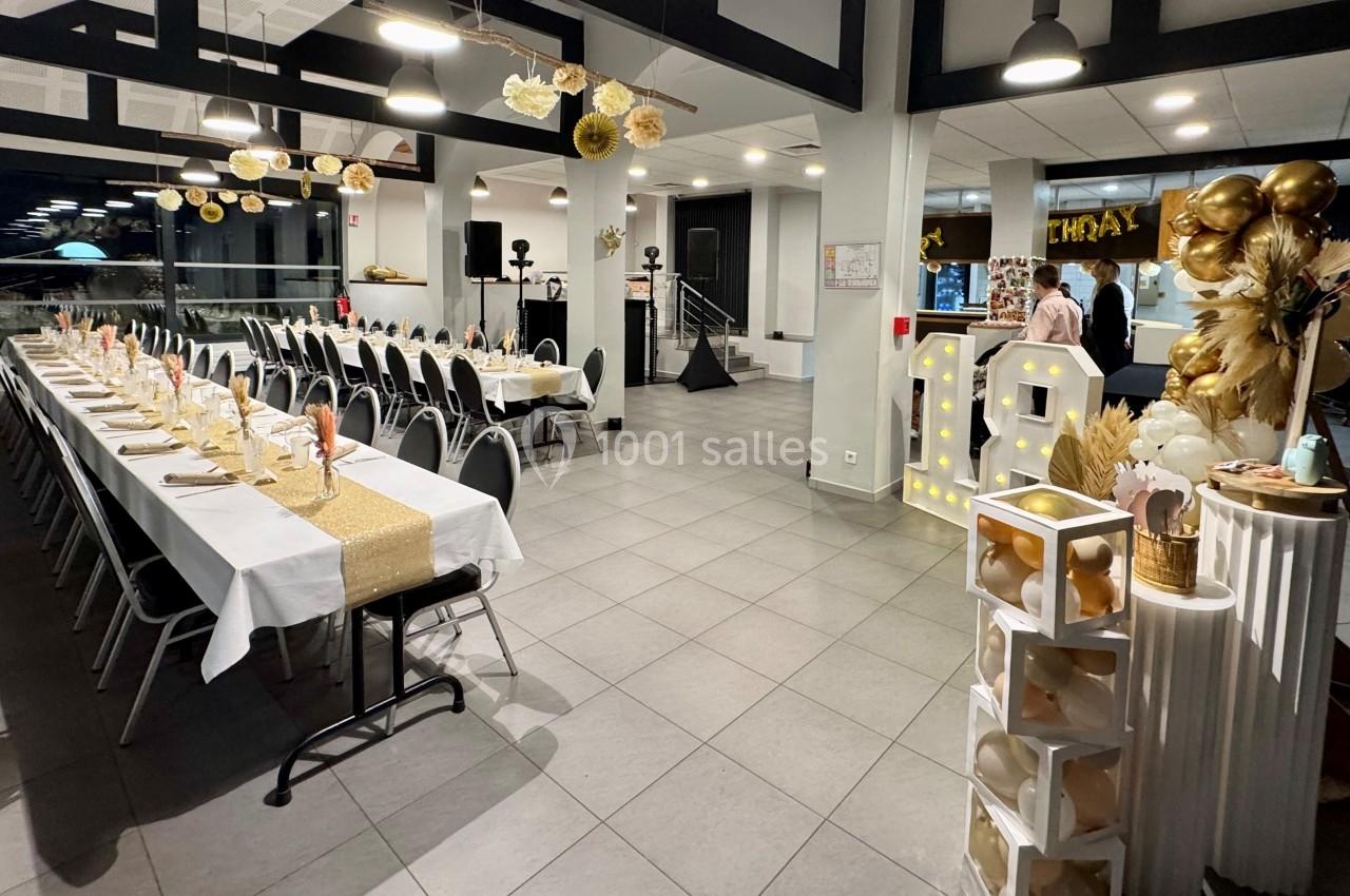 Salle décorée pour une fête, avec longues tables dressées, ballons dorés et blancs, et éléments lumineux.