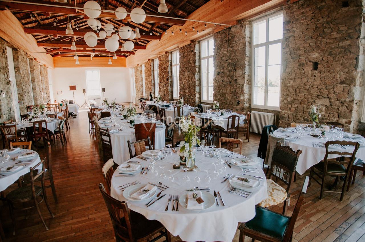 Salle de réception lumineuse avec des tables rondes dressées, entourées de chaises variées, et murs en pierre apparente.