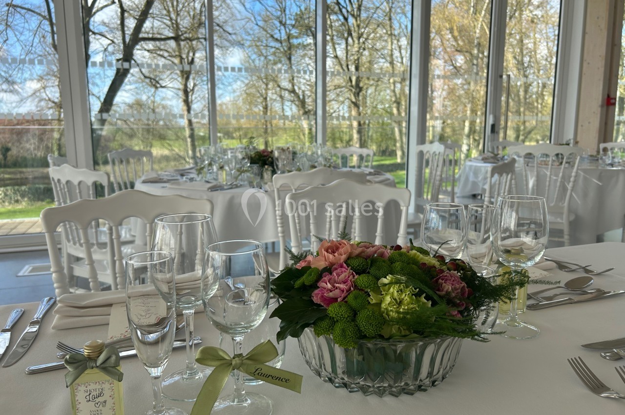 Salle lumineuse avec tables dressées pour un événement, décorées de fleurs et entourées de grandes baies vitrées.