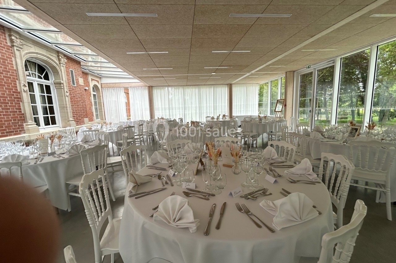 Salle de réception lumineuse avec tables rondes dressées, nappes blanches, serviettes pliées et couverts disposés.