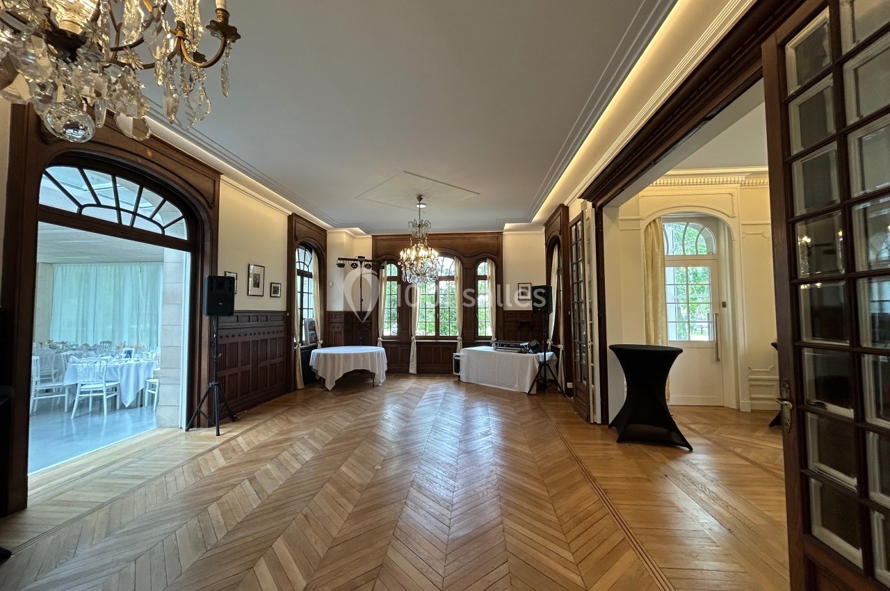 Salle élégante avec parquet en bois, lustres, grandes fenêtres et tables dressées visibles dans une pièce adjacente.