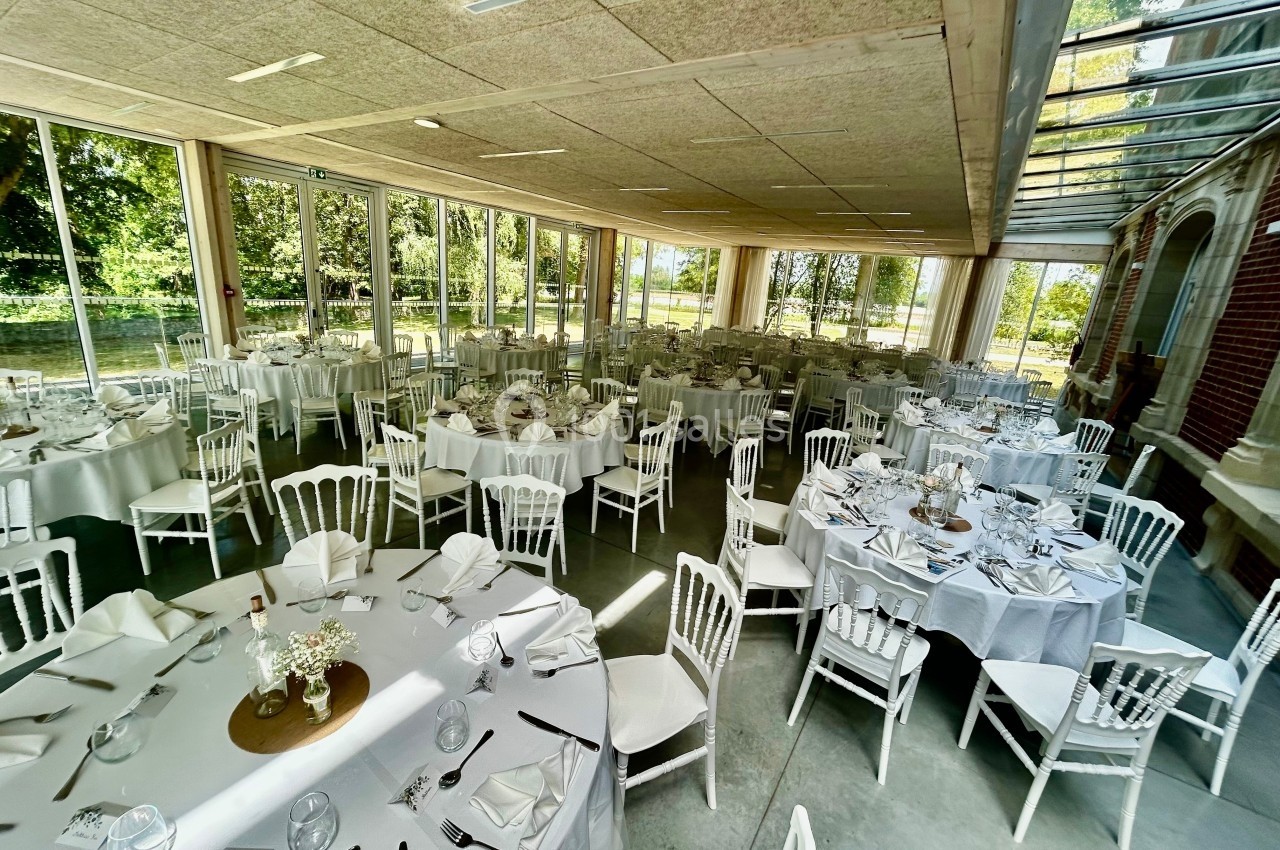 Salle de réception lumineuse avec de grandes baies vitrées, tables rondes dressées et chaises blanches.