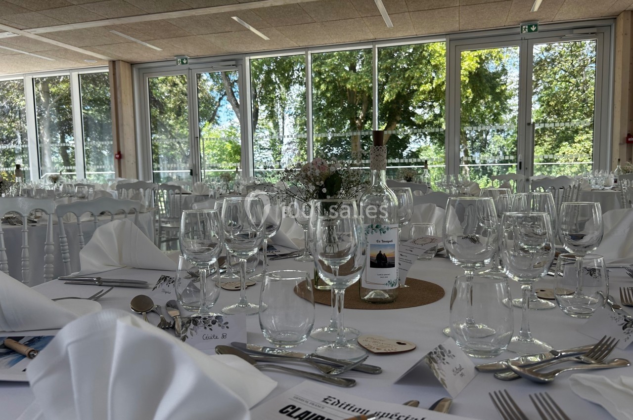 Salle de réception lumineuse avec tables dressées, nappes blanches, verres, couverts et décoration florale.