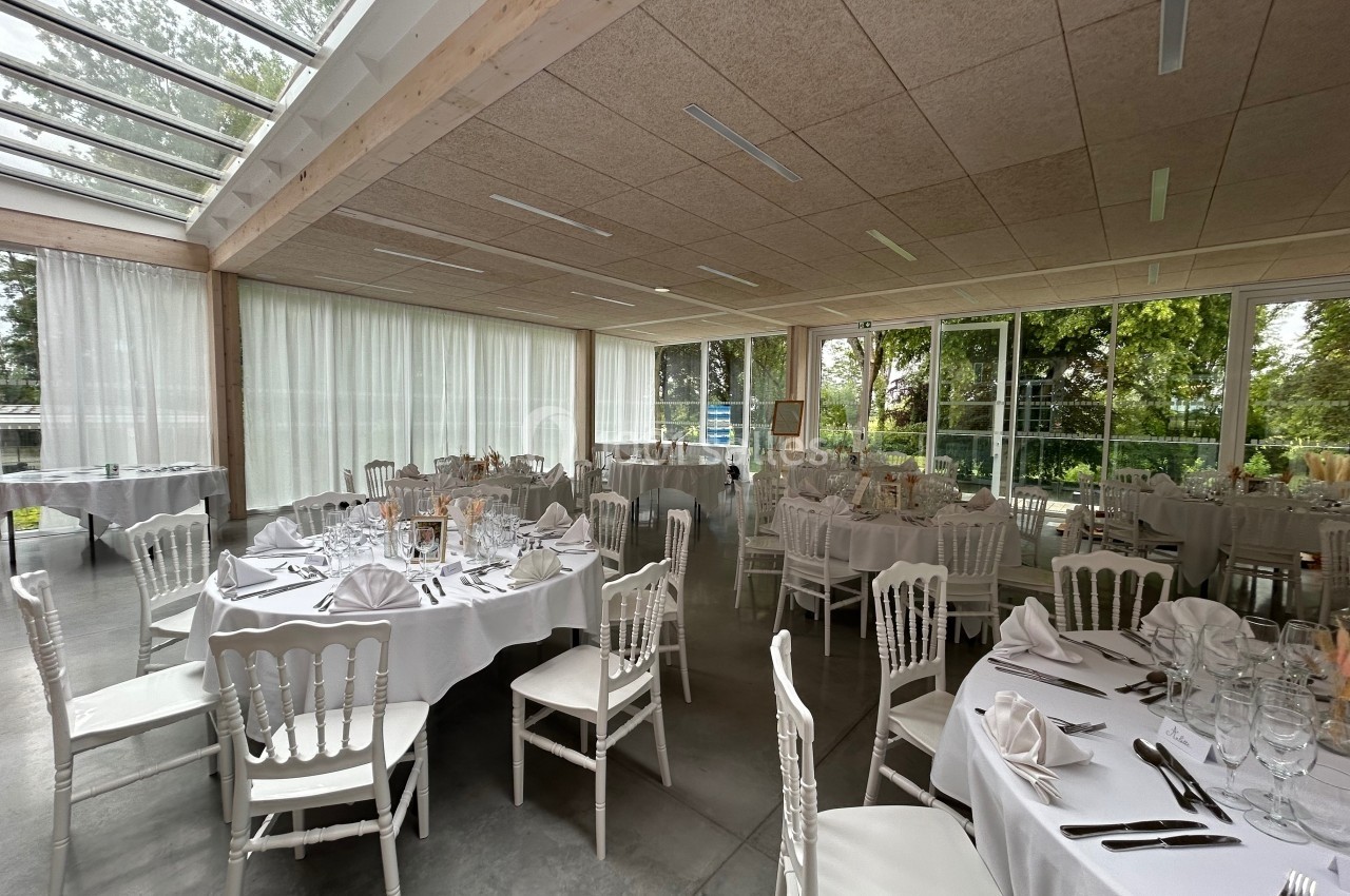 Salle de réception lumineuse avec tables rondes dressées de nappes blanches, entourée de grandes baies vitrées.