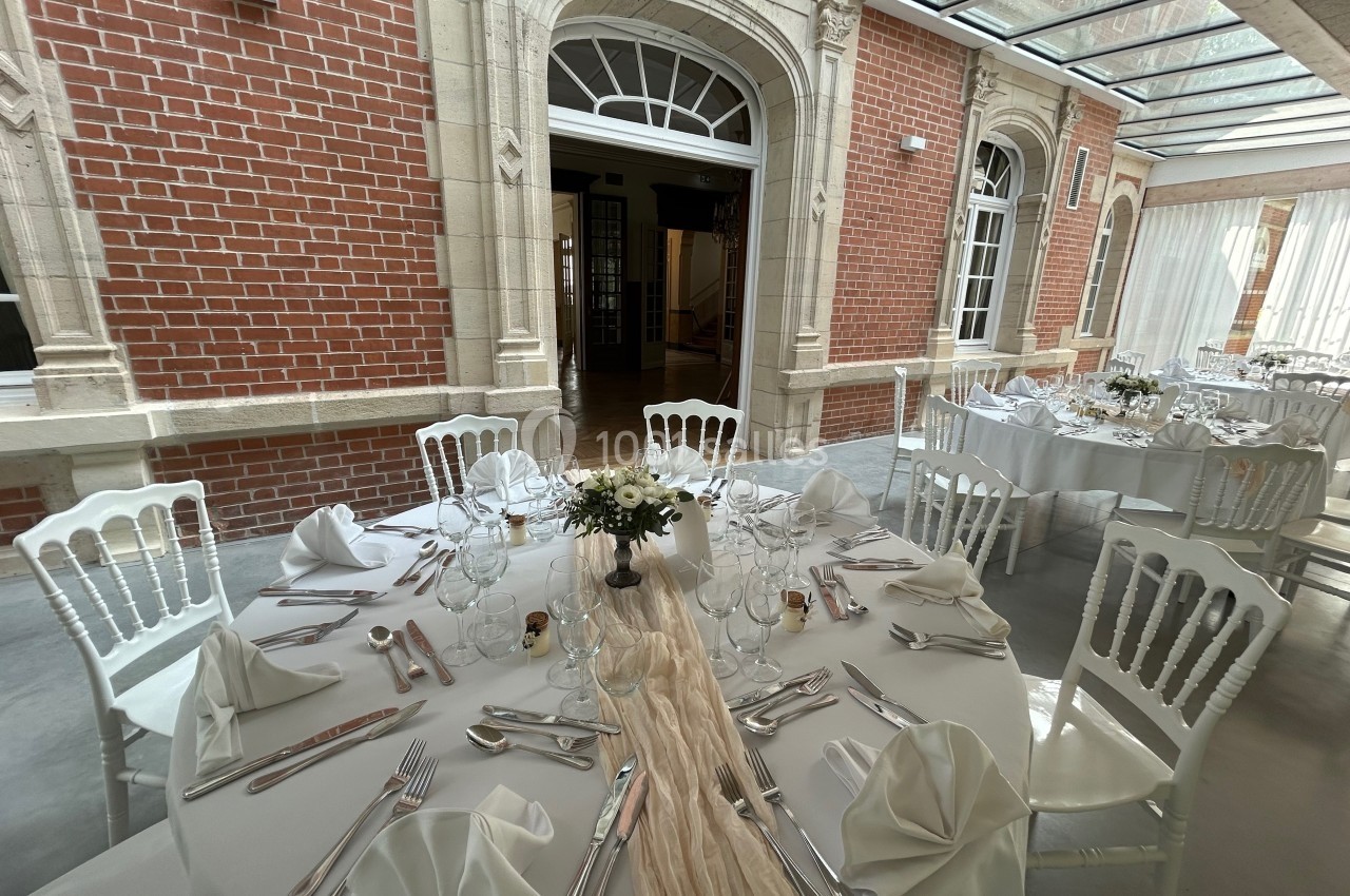 Salle de réception lumineuse avec tables rondes dressées, nappes blanches, chaises élégantes et décoration florale.