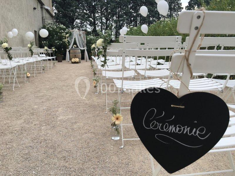 Allée décorée pour une cérémonie en plein air avec des chaises blanches, fleurs et panneaux en ardoise.