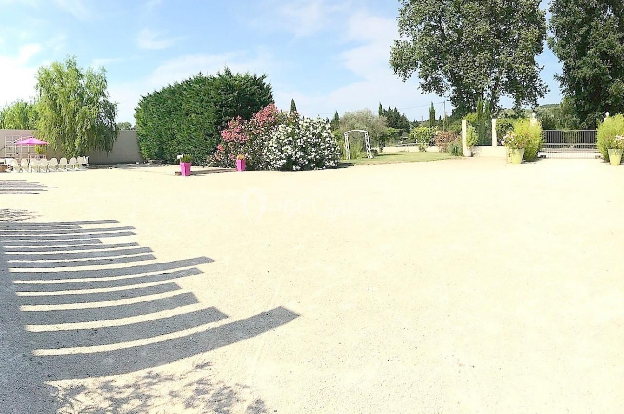 Cour extérieure ensoleillée avec allée gravillonnée, arbres, arbustes fleuris et mobilier de jardin en arrière-plan.