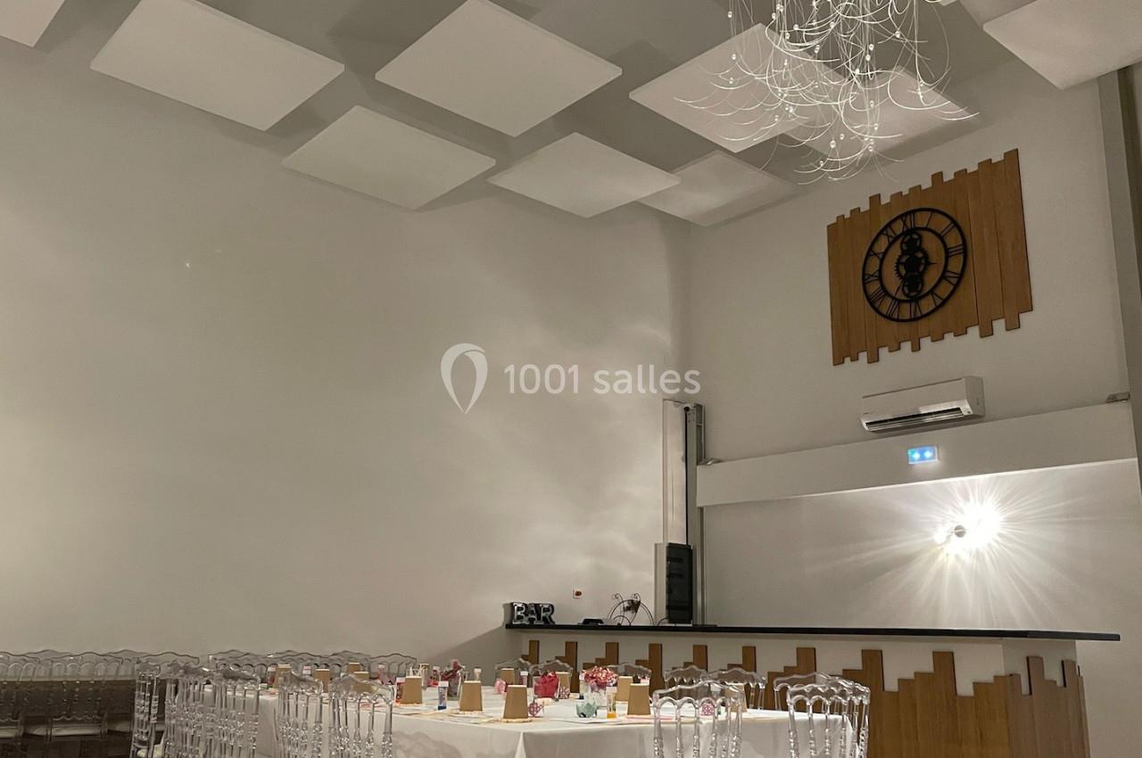 Salle de réception moderne avec tables dressées, chaises transparentes, un bar et une horloge murale décorative.