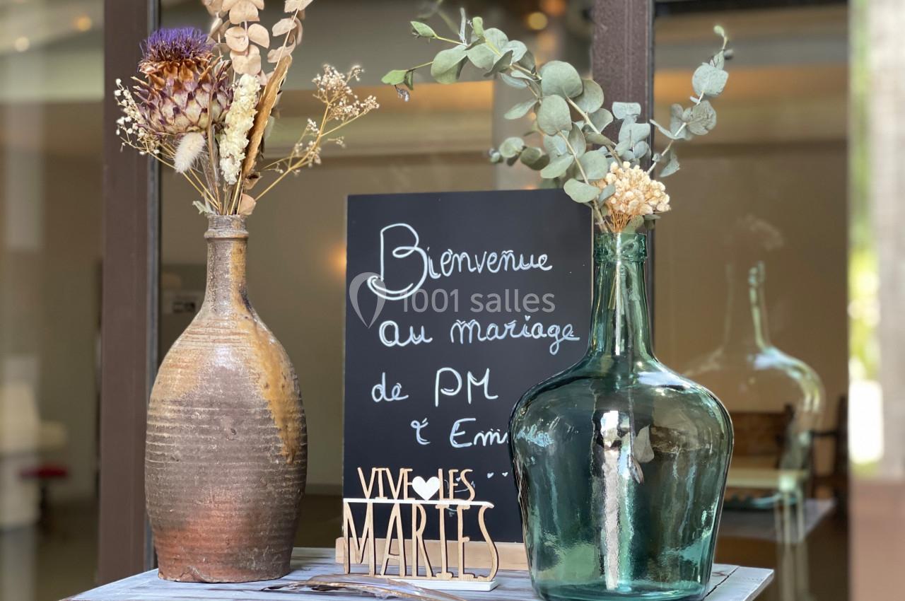 Deux vases avec des fleurs séchées et une ardoise indiquant ’Bienvenue au mariage de PM et Emilie’.