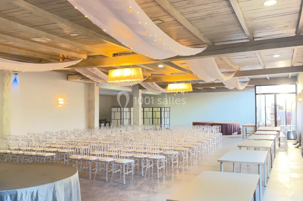 Salle de réception lumineuse avec des chaises transparentes alignées, des tables blanches et des voilages suspendus.