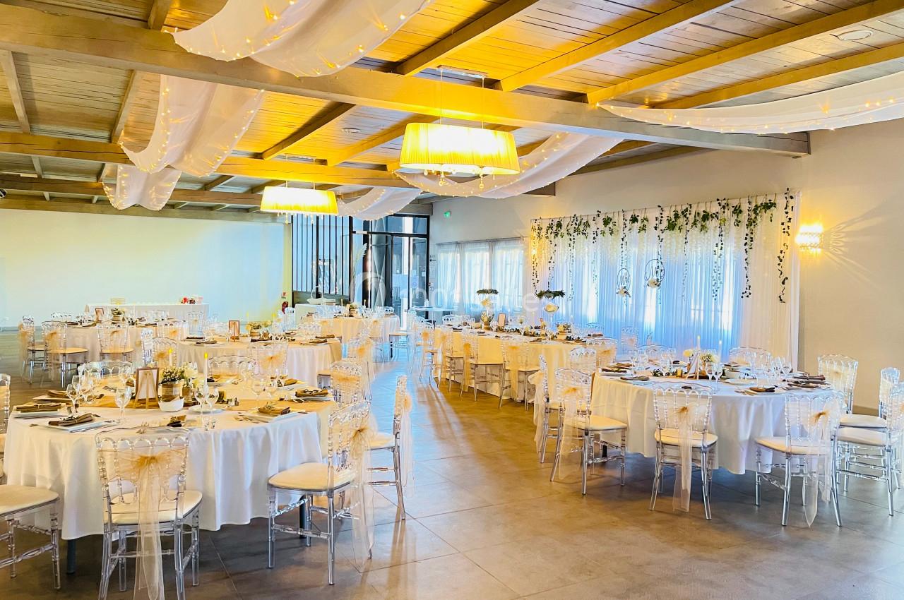 Salle de réception décorée avec des tables rondes dressées, des chaises transparentes et des drapés suspendus au plafond.
