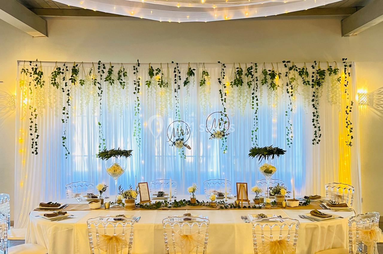Table de réception décorée avec des chaises transparentes, guirlandes lumineuses, feuillages suspendus et fleurs blanches.