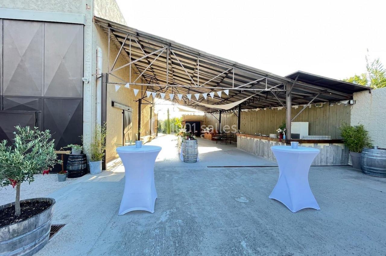 Espace extérieur avec tables hautes drapées de blanc, décoré pour un événement sous une structure métallique ouverte.