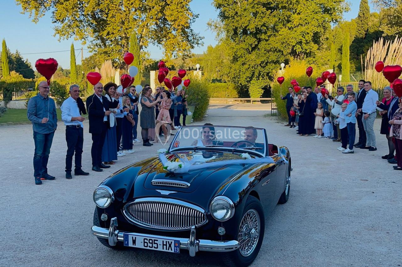Voiture ancienne décapotable noire avec un couple à bord, entourée d'invités tenant des ballons rouges dans un jardin.